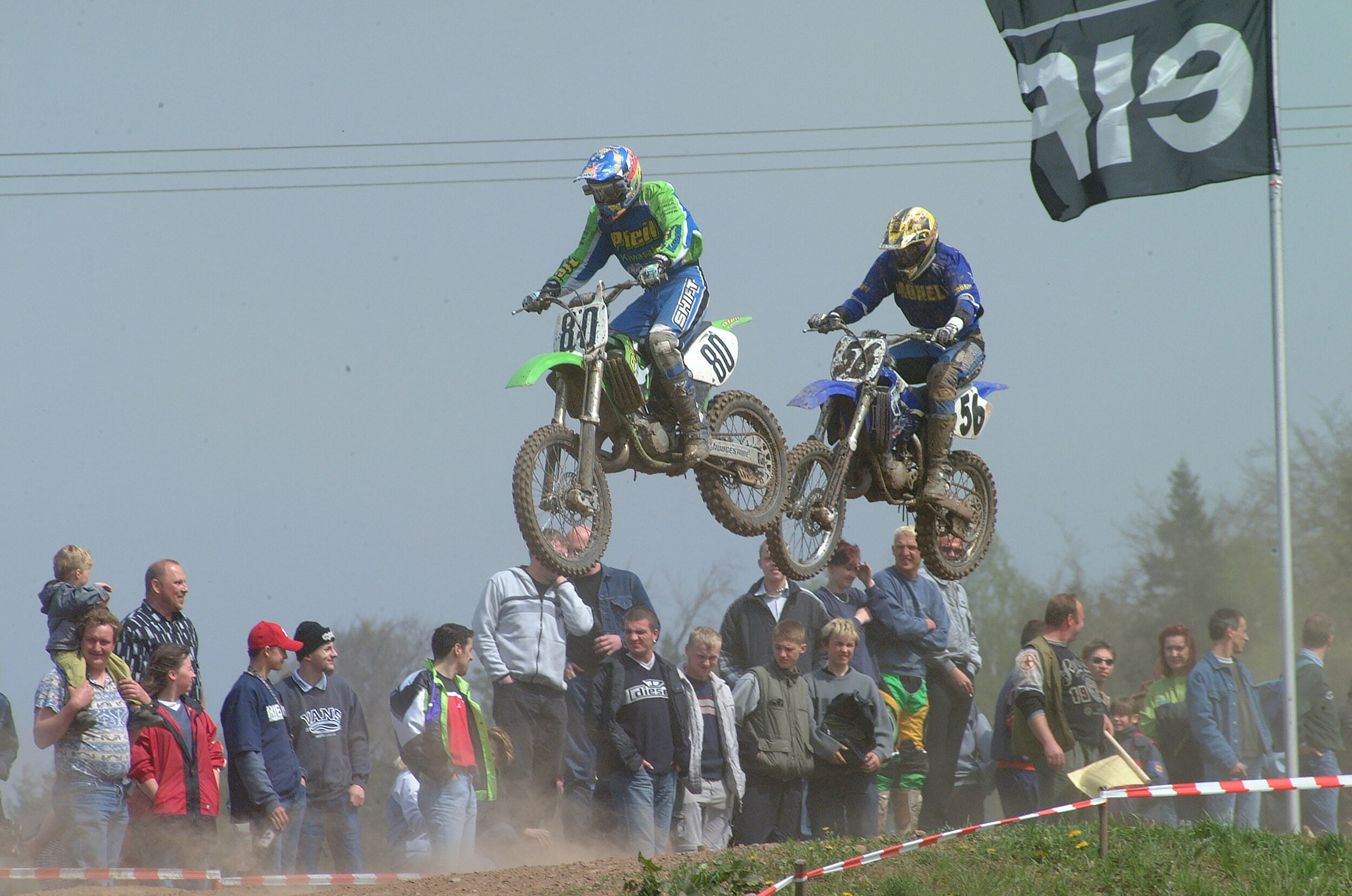 Zwei Motocross-Fahrer fliegen in der Luft über eine staubige Piste, während Zuschauer gespannt zuschauen.
