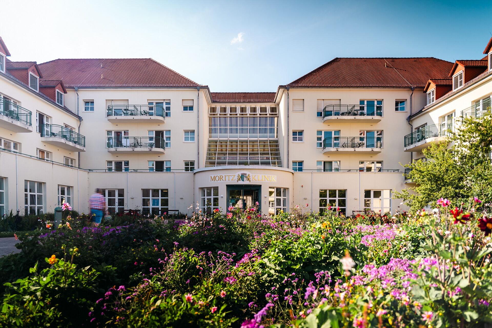 Die Moritzklinik in Bad Klosterlausnitz wird von bunten Blumen im Vordergrund eingerahmt, während eine Person den Eingang passiert.