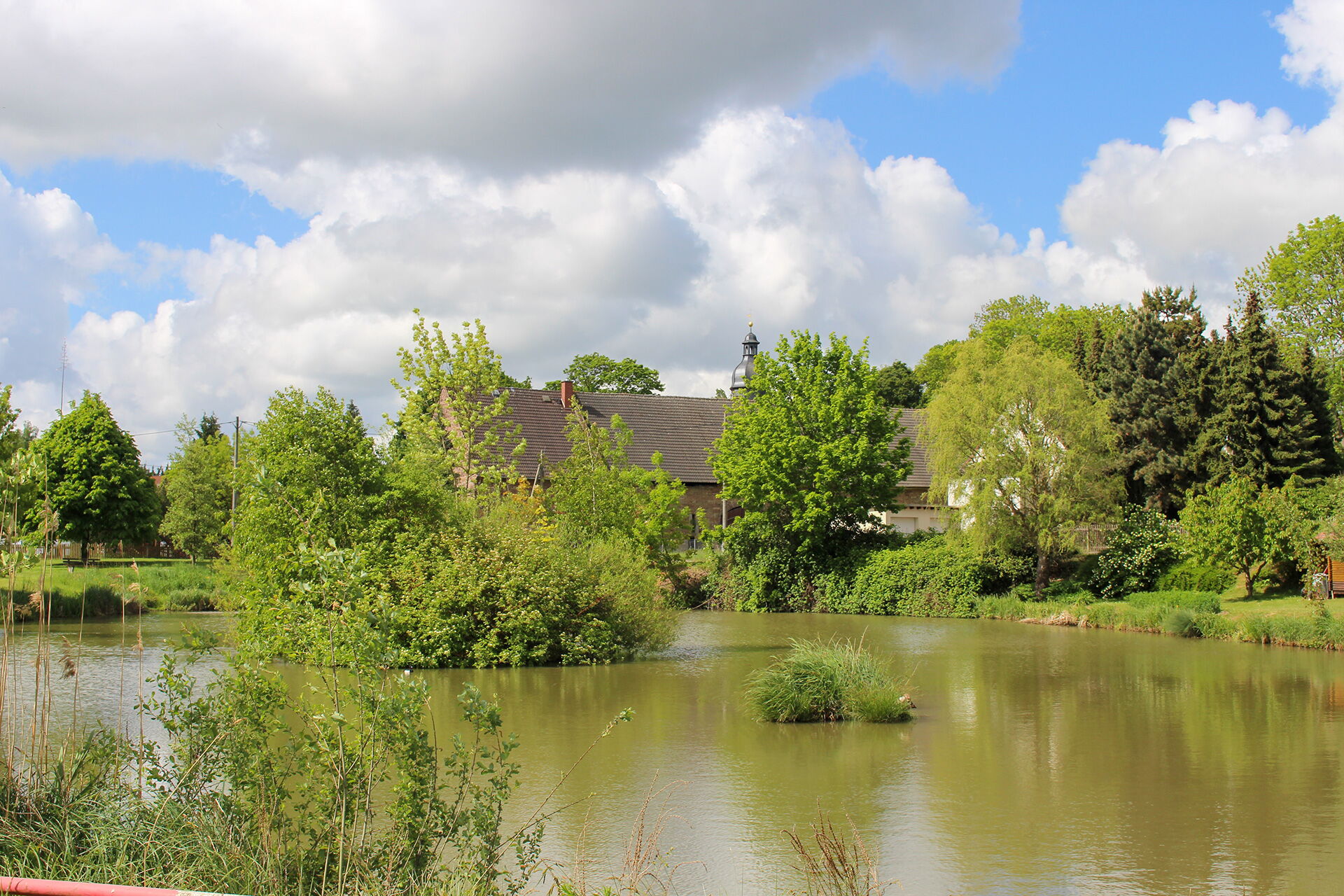 Der Brauteich in Serba spiegelt üppige Bäume und ein Gebäude, unter einem Himmel mit weißen Wolken, wider.