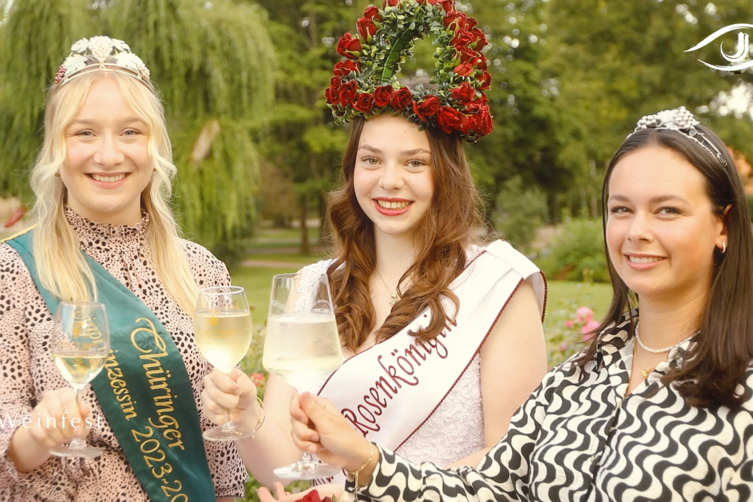 Drei Frauen in festlicher Kleidung stoßen bei einem Weinfest im Freien mit Weingläsern an.