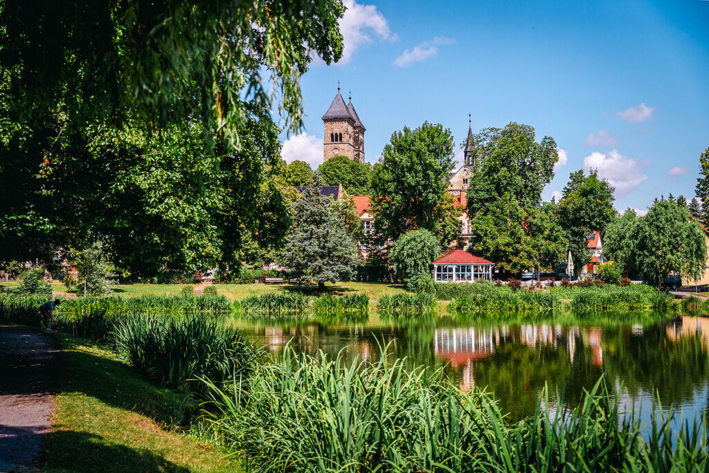 Ein idyllischer Teich in Bad Klosterlausnitz spiegelt Bäume und historische Gebäude unter blauem Himmel wider.