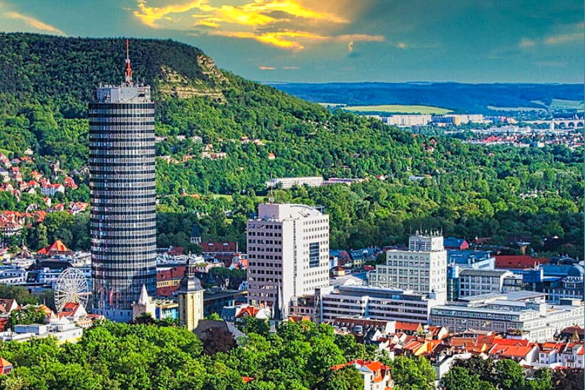 Der markante JenTower erhebt sich über die grüne Stadtlandschaft von Jena bei Sonnenuntergang, umgeben von Hügeln und moderner Architektur.