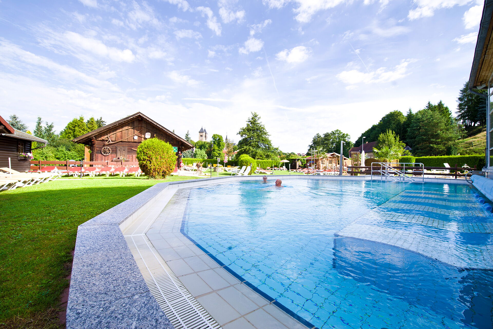 Ein kristallklarer Pool in einem idyllischen Garten mit Sauna-Holzhäusern lädt zum Entspannen in der Kristall Therme ein.