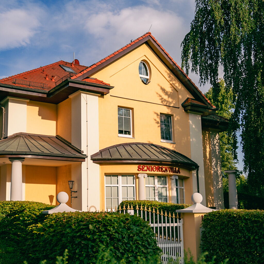 Eine gelbe Seniorenresidenz mit rotem Dach und gepflegtem Garten strahlt im Sonnenlicht.