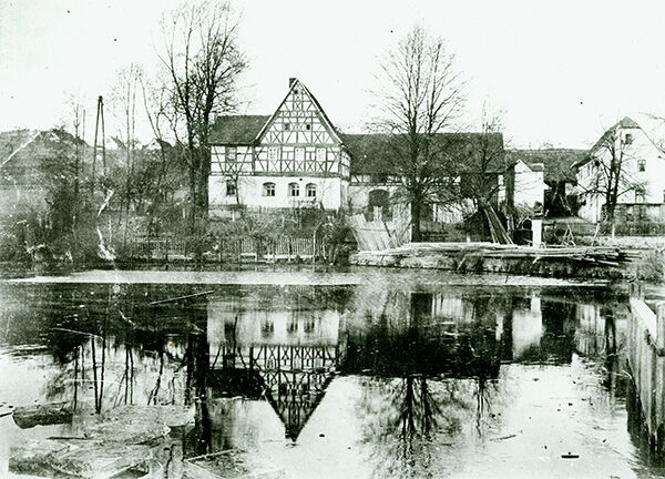 Historische Fachwerkhäuser spiegeln sich in einem ruhigen Teich im frühen 20. Jahrhundert wider.