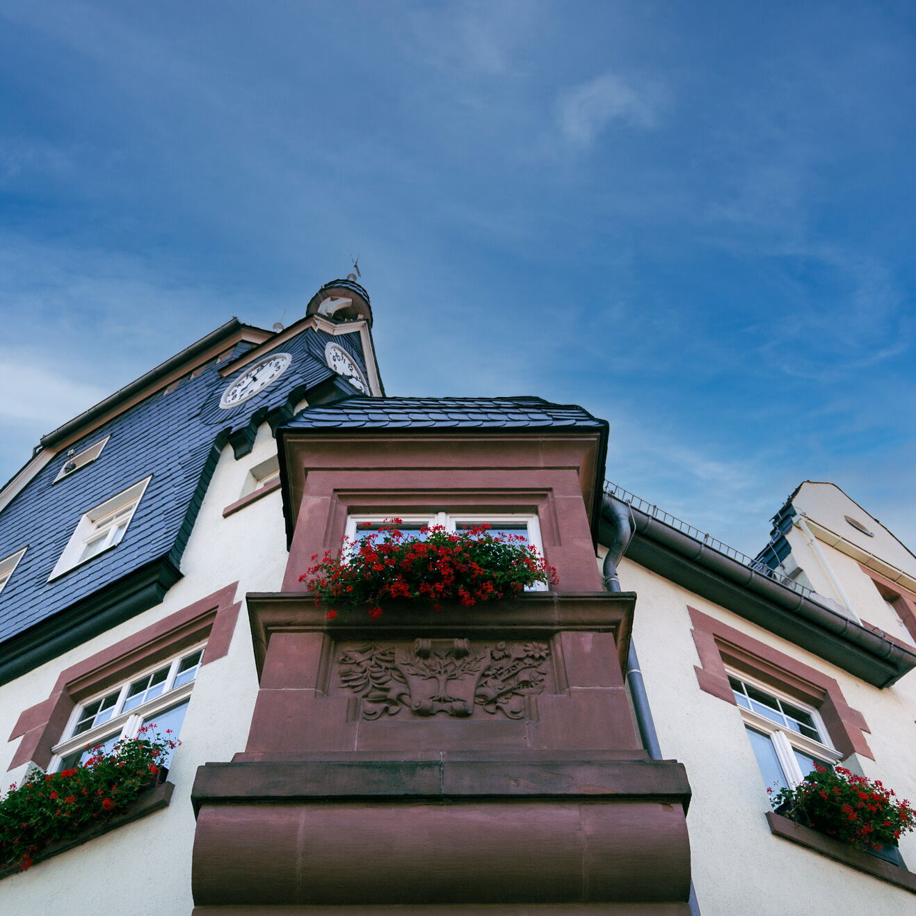 Das historische Rathaus mit blühenden Geranien und einer Turmuhr erhebt sich vor klarem blauen Himmel.
