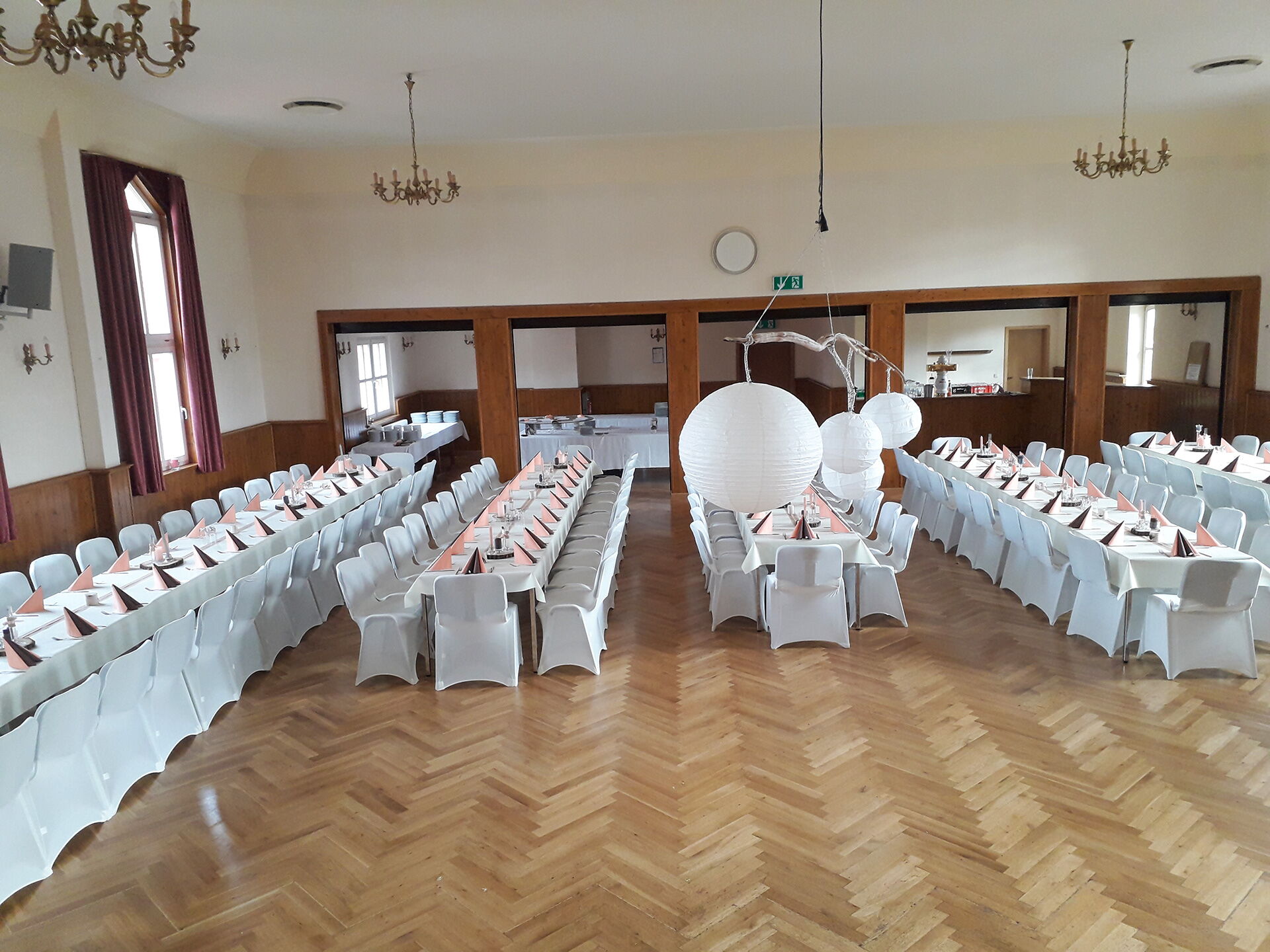 Ein großer Saal ist mit weiß gedeckten Tischen und Stühlen für ein elegantes Event vorbereitet, mit dekorativen Lampions.