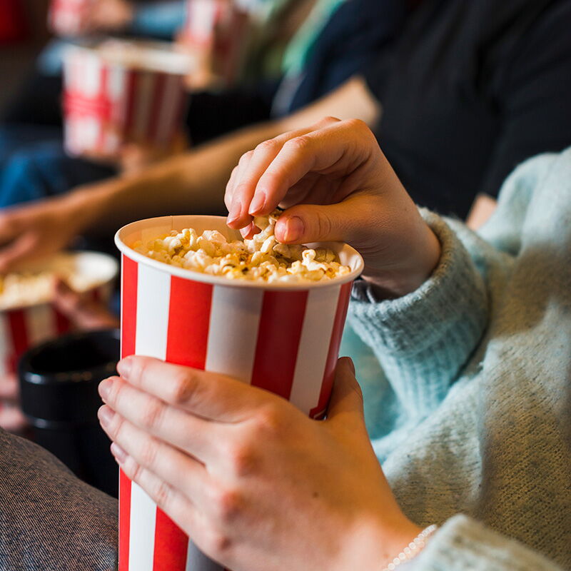 Eine Person greift in einen rot-weiß gestreiften Popcornbecher im Holzlandkino, umgeben von anderen Zuschauern.