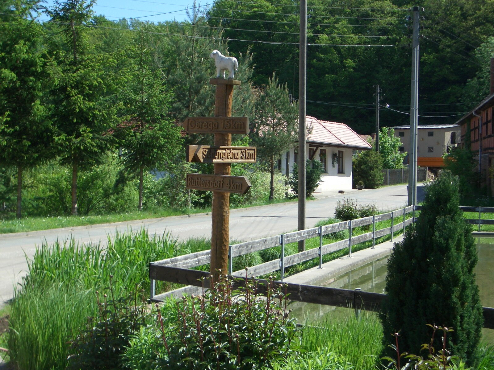 Ein hölzerner Wegweiser mit Schafskulptur zeigt Entfernungen in einer grünen, ländlichen Umgebung an.