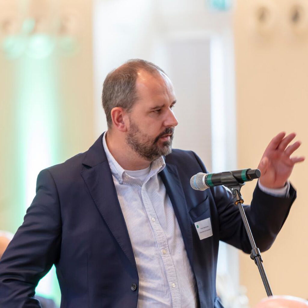 Ein Mann im Anzug hält eine Rede vor einem Mikrofon in einem eleganten Raum, möglicherweise als Bürgermeister.