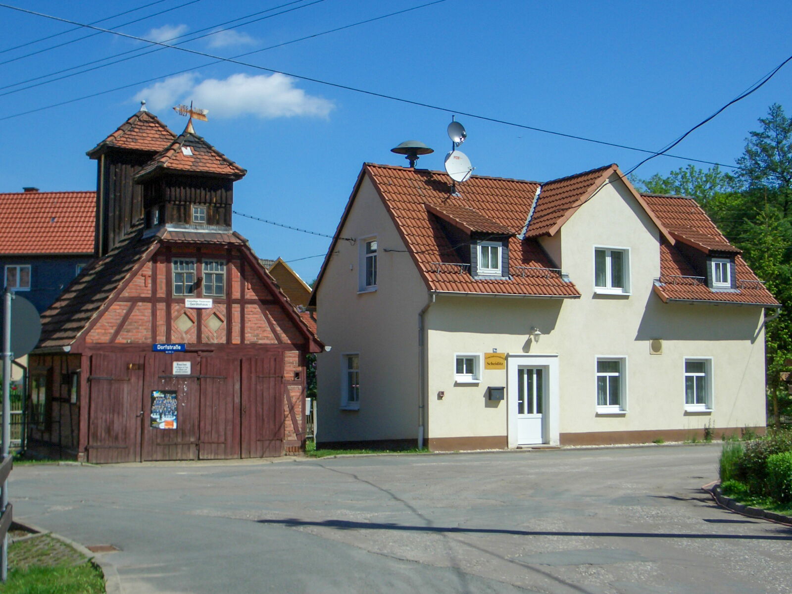 Zwei traditionelle Gebäude, ein Fachwerkhaus und das Gemeindezentrum in Scheiditz, stehen unter blauem Himmel in einer ruhigen Straße.