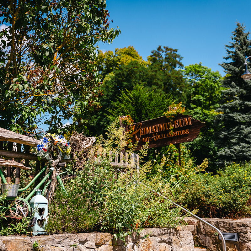 Ein Schild des Heimatmuseums Bad Klosterlausnitz steht umgeben von üppigem Grün und einem dekorativen Fahrrad.