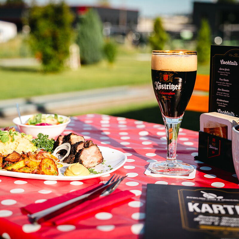 Ein Teller mit gegrilltem Fleisch und Beilagen steht auf einem rot-weiß gepunkteten Tisch neben einem Glas dunklem Bier im Gasthaus am Kurpark.
