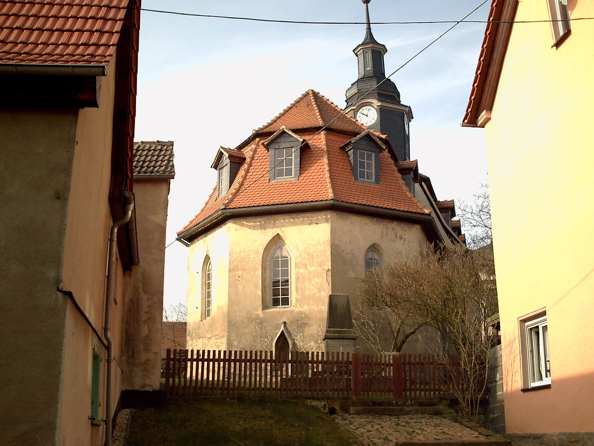 Eine malerische Kirche mit rotem Ziegeldach steht zwischen zwei Gebäuden in einem idyllischen Dorf.
