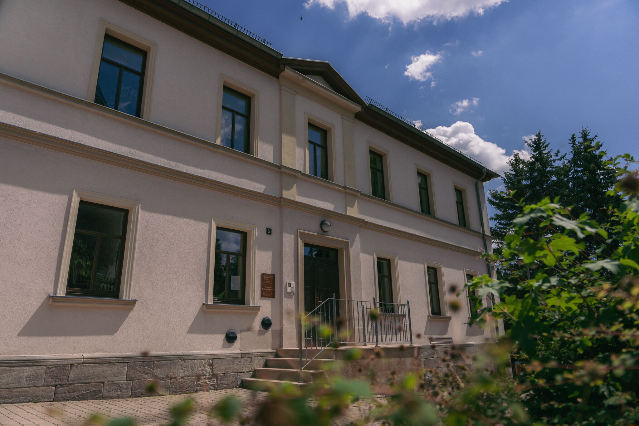 Ein zweistöckiges Gebäude mit weißer Fassade und mehreren Fenstern steht unter einem blauen Himmel mit Wolken.