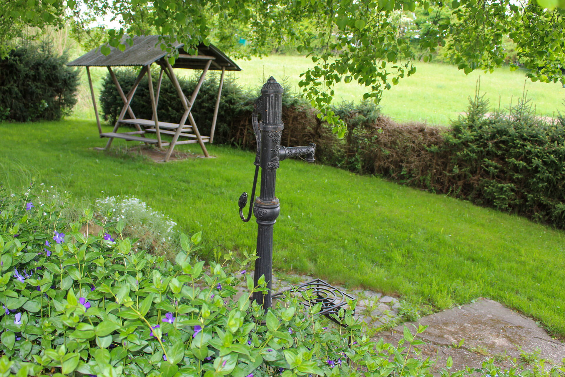 Eine alte Wasserpumpe steht in einem grünen Garten neben einer Holzbank, umgeben von üppigem Laub in der Gemeinde Serba.