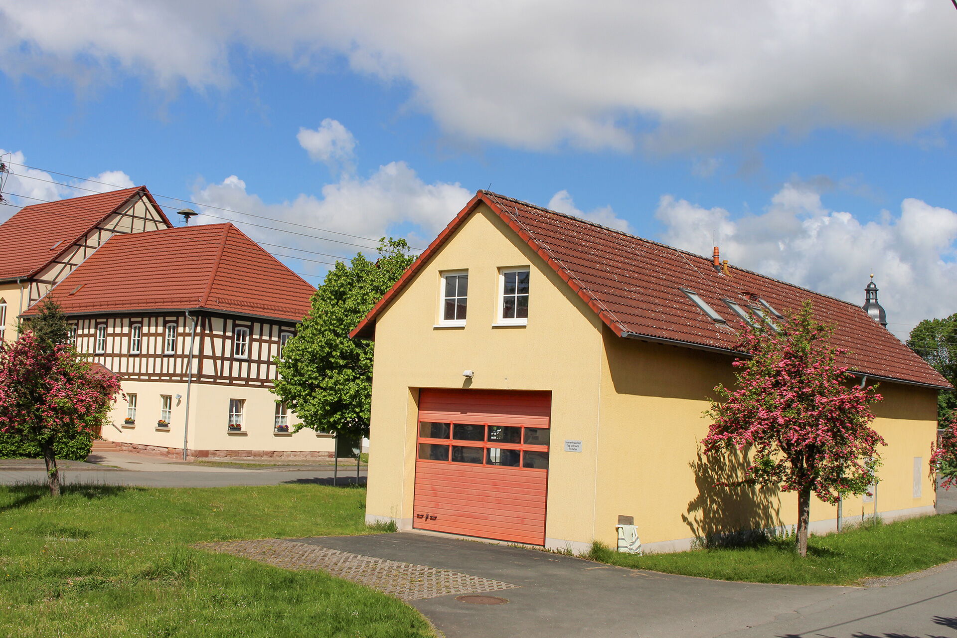 Ein Feuerwehrhaus in Serba steht neben traditionellen Fachwerkhäusern unter einem blauen Himmel mit weißen Wolken.
