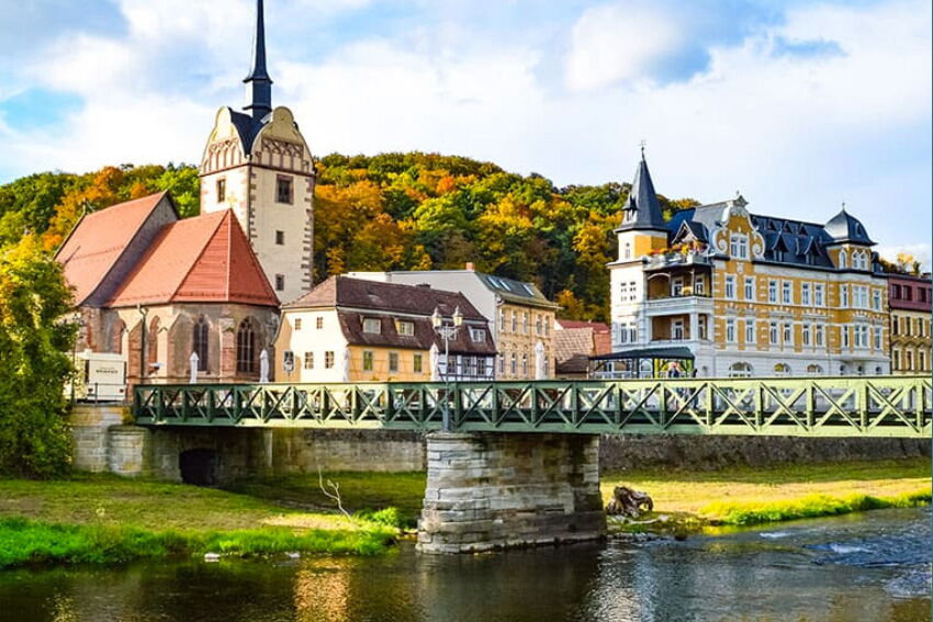 Eine malerische Brücke in Gera überquert einen Fluss vor historischen Gebäuden und einem herbstlich bunten Wald.
