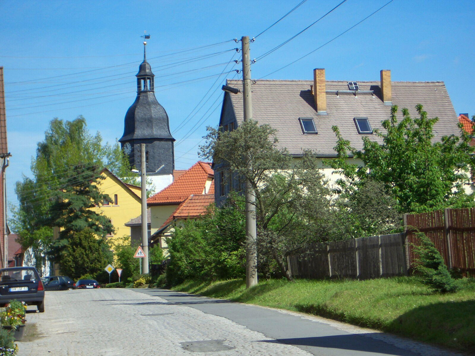 Eine ruhige Dorfstraße in Albersdorf mit markantem Kirchturm und gepflegten Häusern, die von Bäumen umgeben sind.