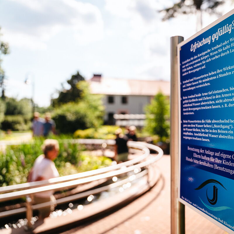 Ein Schild erklärt die Kneippanlage in Bad Klosterlausnitz, während Menschen im Hintergrund die Anlage genießen.