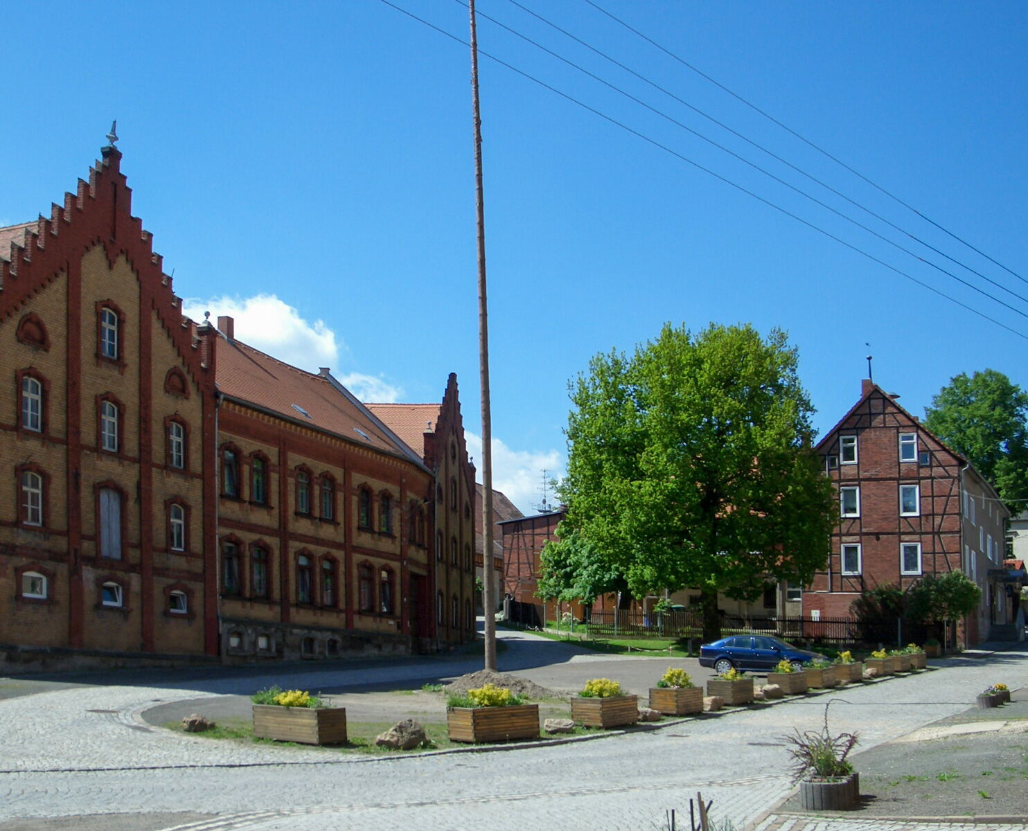 Dorfplatz in Bobeck mit historischen Backsteingebäuden, grünen Bäumen und einem geparkten blauen Auto unter klarem Himmel.