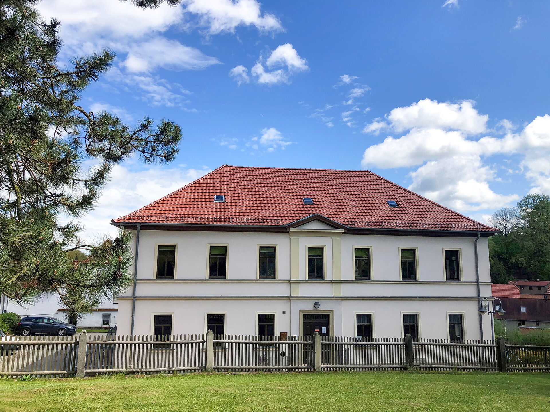 Ein weißes Gebäude mit rotem Dach und Zaun steht unter blauem Himmel in einem grünen Gemeindebereich.