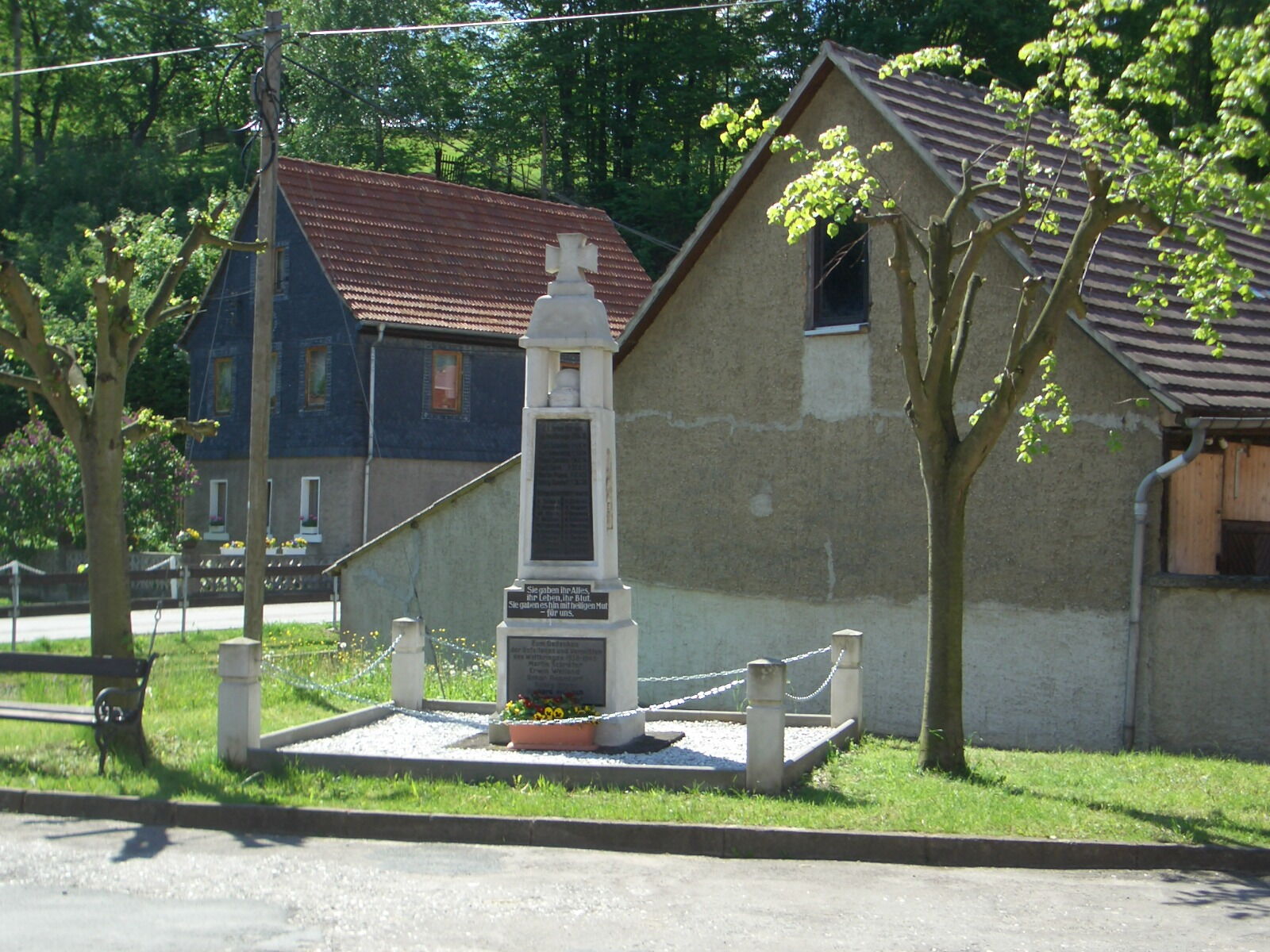 Ein Kriegerdenkmal steht vor rustikalen Häusern, umgeben von blühenden Bäumen und einer sonnigen Landschaft.