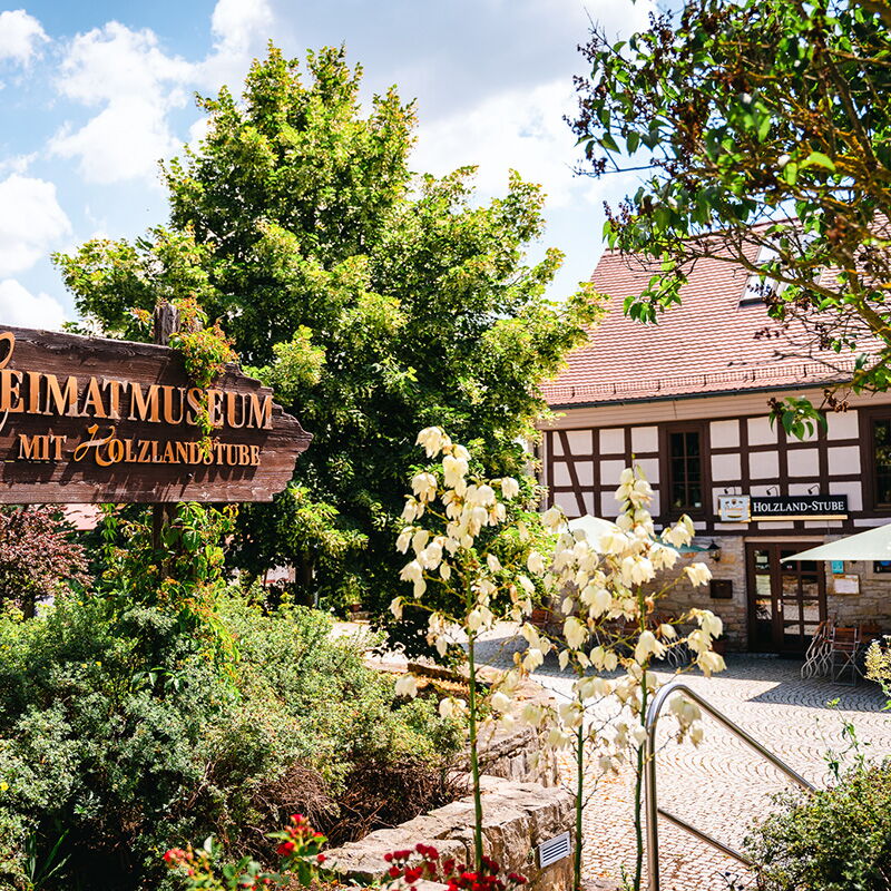 Das Heimatmuseum mit der Holzlandstube in Bad Klosterlausnitz ist von grünen Bäumen und einem blühenden Garten umgeben.
