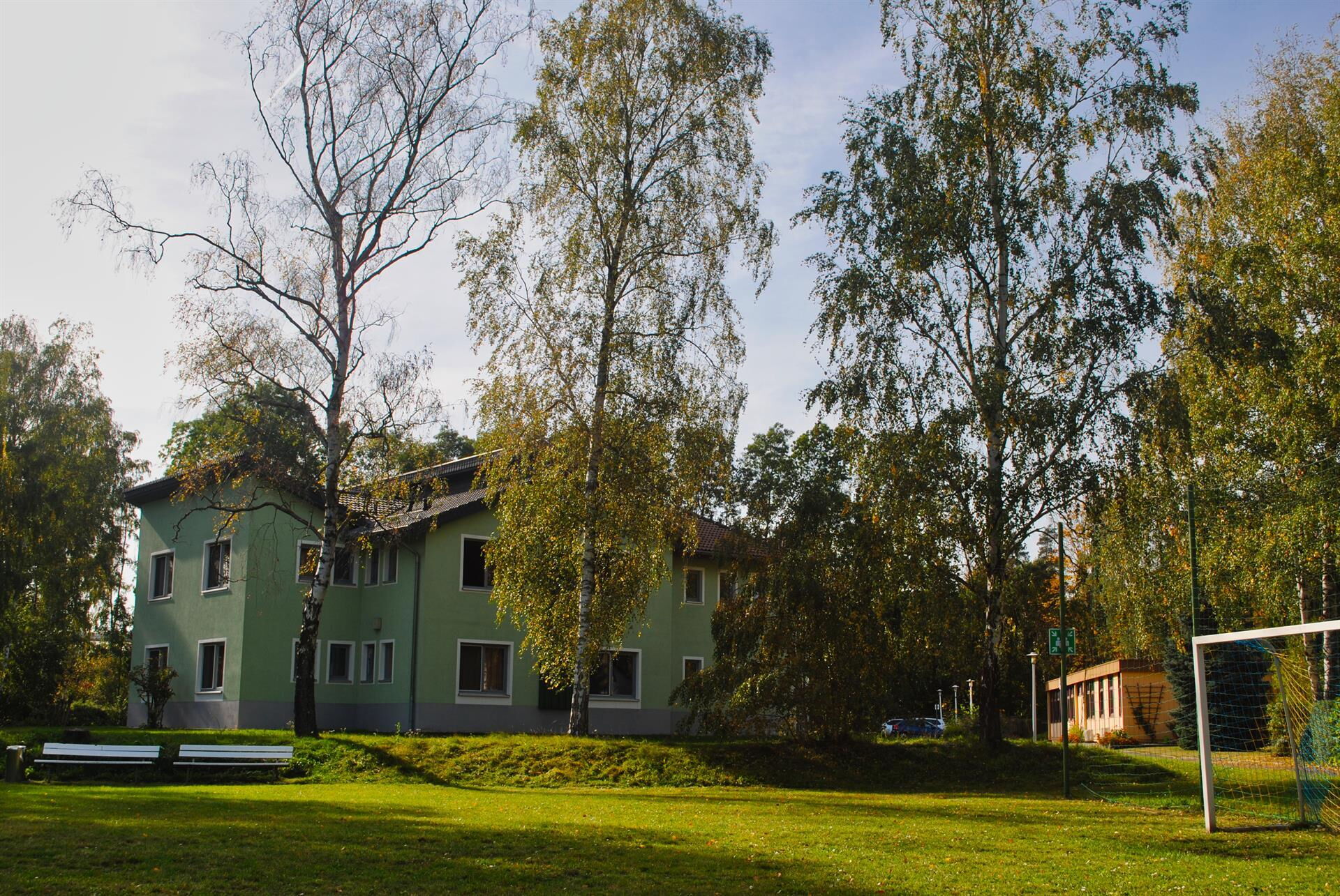 Ein grünes Haus steht umgeben von hohen Bäumen auf einer sonnigen Wiese mit Fußballtor und Bänken.