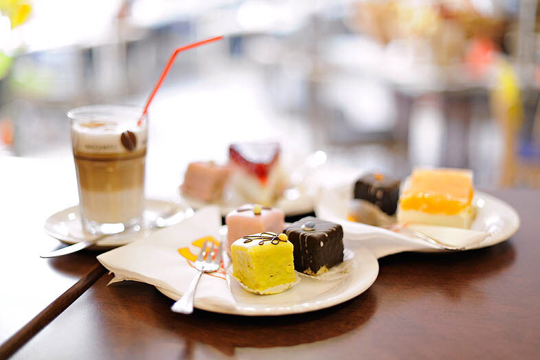 Bunte Petit Fours und ein Glas Latte Macchiato mit Strohhalm stehen einladend auf einem Tisch im Café des Hotel Fabrice.