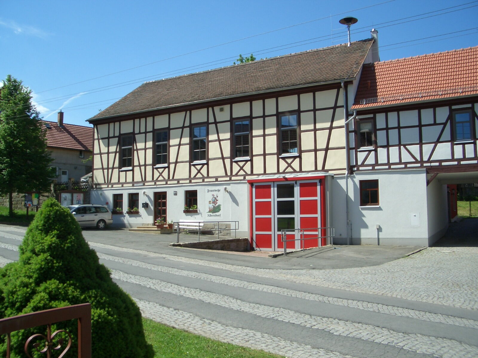 Ein traditionelles Fachwerkhaus mit rotem Tor dient als Gemeindezentrum an einer gepflasterten Straße.