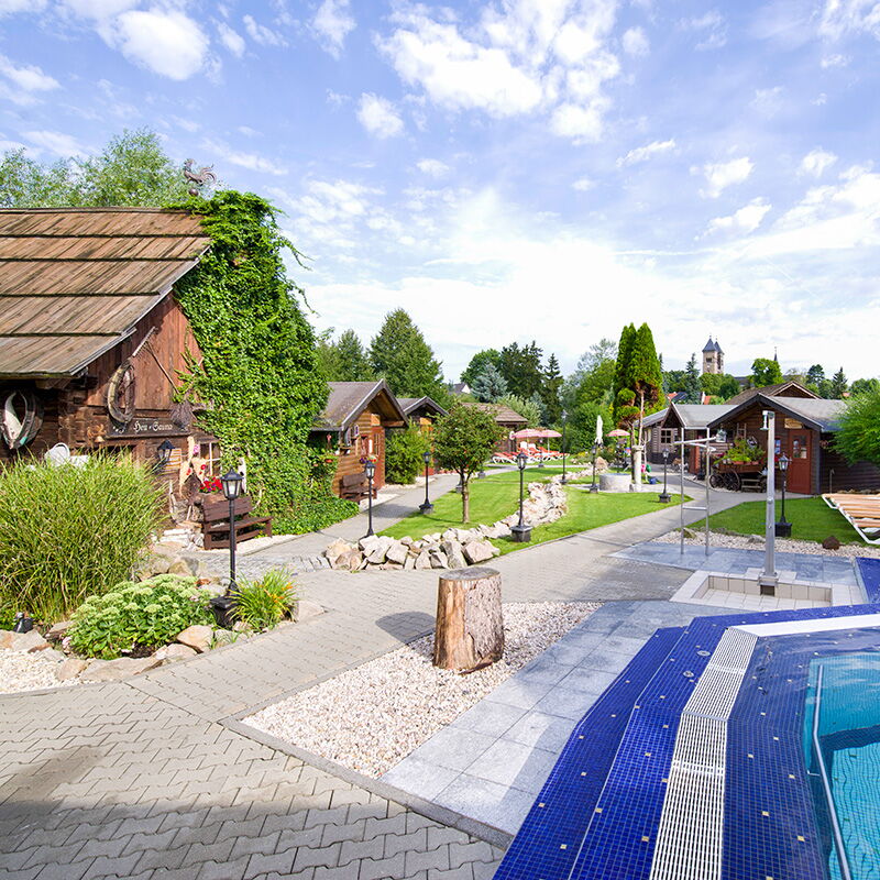 Die Kristall Therme Bad Klosterlausnitz zeigt eine idyllische Außenanlage mit Holzgebäuden und einem blauen Pool.