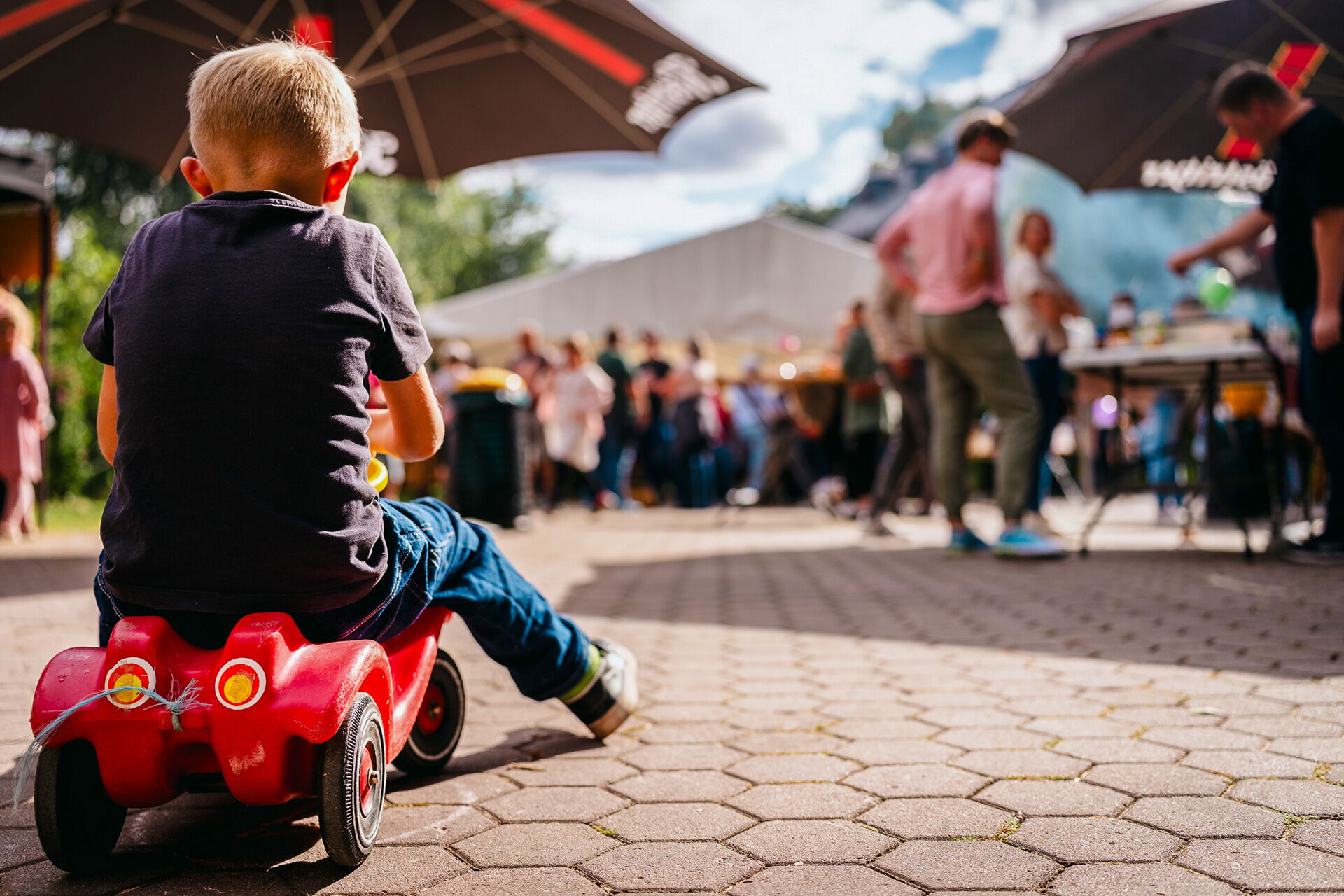 Ein Kind fährt auf einem roten Spielauto bei einem lebhaften Freizeitangebot mit vielen Menschen im Hintergrund.