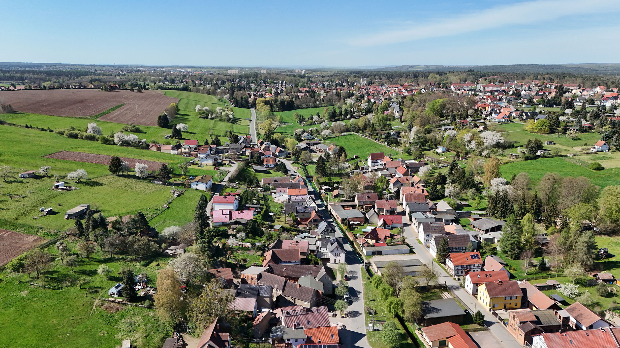 Luftaufnahme eines Dorfes mit grünen Feldern und einer Kirche am Horizont bei strahlend blauem Himmel.