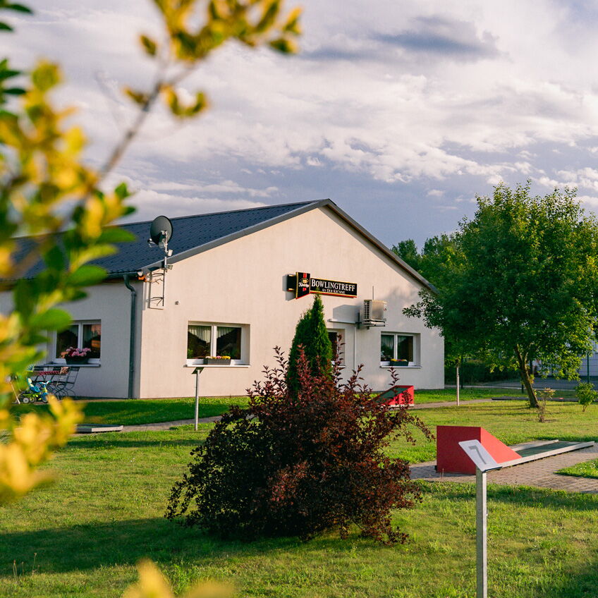 Ein weißes Gebäude mit der Aufschrift "Bowlingtreff" steht auf einer gepflegten, grünen Wiese unter einem bewölkten Himmel.