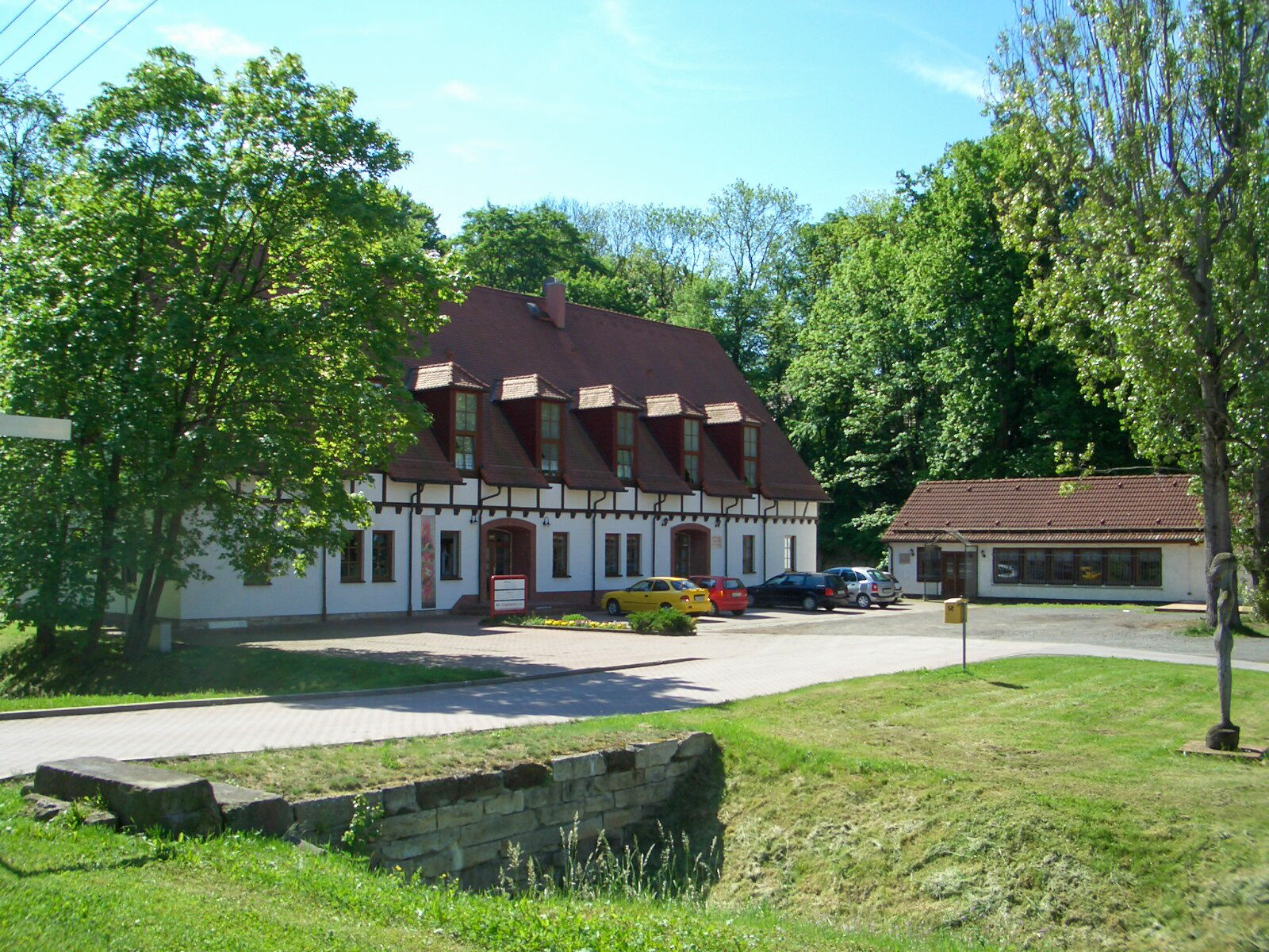 Das ehemalige Konsumgebäude in Schlöben steht umgeben von grünen Bäumen und parkenden Autos unter einem klaren Himmel.