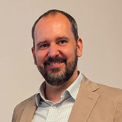 Ein lächelnder Mann in einem beigen Sakko steht vor einem neutralen Hintergrund.