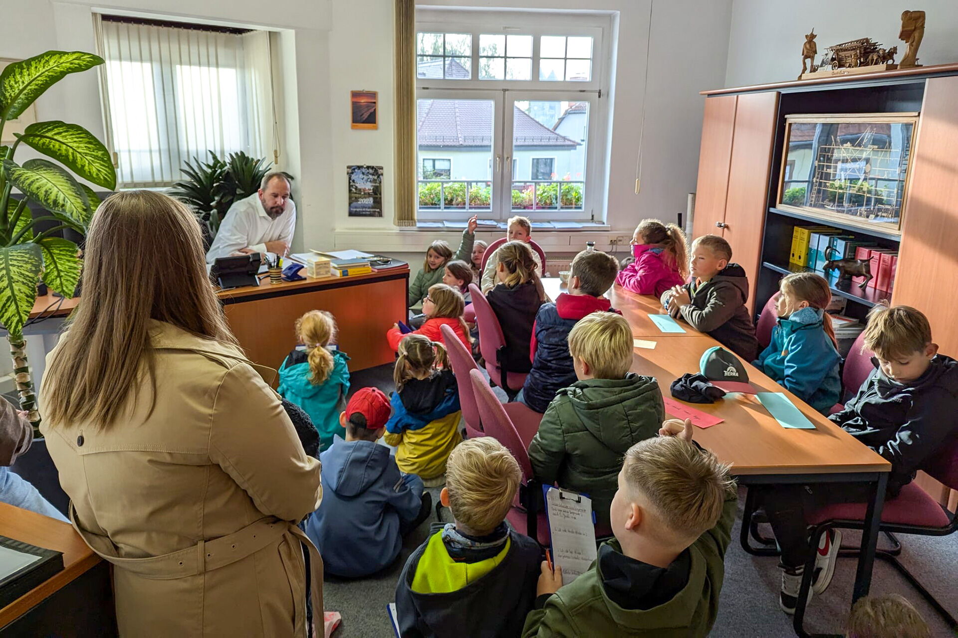 Eine Gruppe von Kindern sitzt aufmerksam in einem Büro und hört einem Mann am Schreibtisch zu.